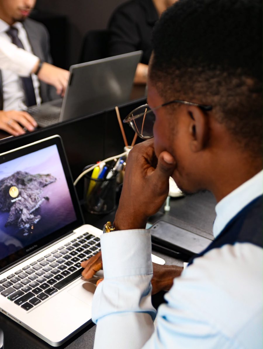 Profissional de camisa azul-clara trabalhando em um notebook em um escritório, com equipe ao fundo